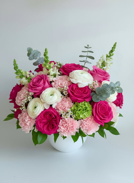 A floral arrangement featuring pink hydrangeas, vibrant hot pink roses, creamy white ranunculus, green hydrangeas, and eucalyptus leaves in a white ceramic container.